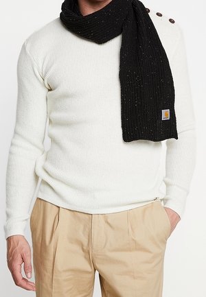 Homme portant un pull crème côtelé avec des boutons aux épaules, un pantalon beige et une écharpe noire tricotée avec un petit écusson logo.