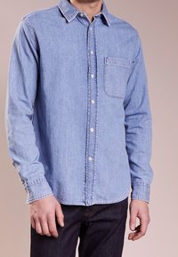 Ljusblå denimskjorta med långa ärmar, knäppning med krage, frampocket och vita knappar. Tyget har en något sliten textur.