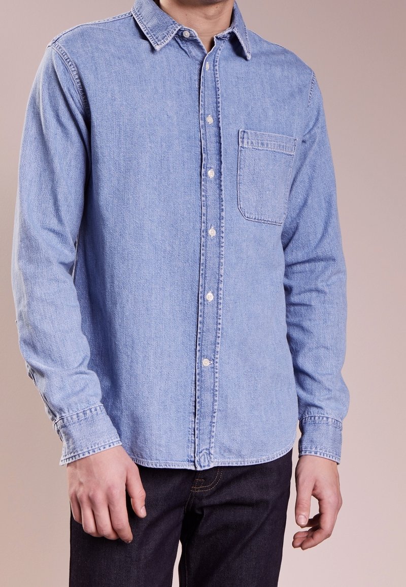Ljusblå denimskjorta med långa ärmar, knäppning med krage, frampocket och vita knappar. Tyget har en något sliten textur.