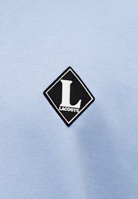 Sweatshirt en coton bleu clair avec un patch en forme de losange noir sur lequel sont brodées en blanc la lettre "L" et "LACOSTE". Texture lisse.