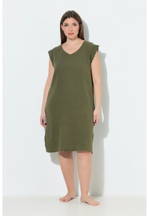 Frau steht barfuß und trägt ein olivgrünes, ärmelloses knielanges Kleid mit Spitzendetails an den Schultern vor weißem Hintergrund.