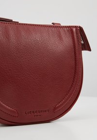 Czerwona skórzana torba na ramię o zaokrąglonym kształcie, teksturowanej powierzchni i tłoczonym logo. Posiada zamek błyskawiczny i subtelne detale przeszyć.