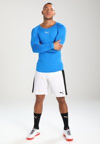 Maglietta sportiva blu a maniche lunghe, pantaloni chiari con strisce nere, calze nere fino al ginocchio e scarpe sportive bianche con dettagli rossi.