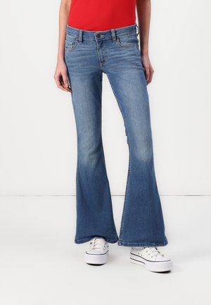 Uitlopende blauwe denim jeans met een medium taille, lichte vervaging, vijf-pocket ontwerp en stikdetails; gedragen met witte sneakers.