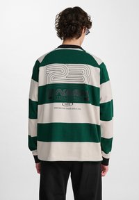 Långärmad rugbytröja i grön och krämrandig design. Har stor tryckt text på baksidan, svart krage och kontrasterande muddar.