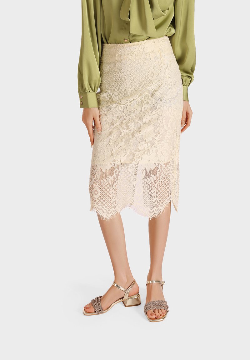 Gonna a matita in pizzo beige chiaro, fino al ginocchio, con orlo scallopato e motivi floreali a strati, abbinata a una blusa verde e sandali metallici con cinturini.