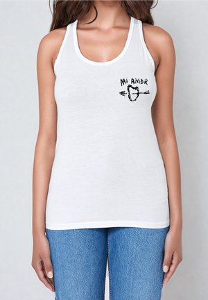 Nainen pukeutuneena valkoiseen toppiin, jossa vasemmassa rinnassa "Mi Amor" -teksti ja nuoli-sydän-kuvio, yhdistettynä sinisiin farkkuihin, seisomassa yksinkertaisen taustan edessä.