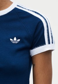 Gros plan du haut du bras d'une personne portant un t-shirt Adidas bleu marine avec un liseré blanc et un détail à trois bandes blanches sur la manche.