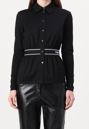 Vrouw draagt een zwart overhemd met lange mouwen en knopen, met een gestreepte elastische tailleband en zwarte leren broek, staand tegen een witte achtergrond.