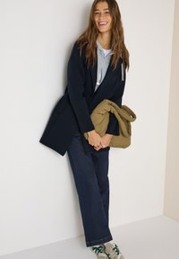 Femme souriante portant un manteau bleu marine par-dessus un sweat à capuche gris et un jean, tenant une doudoune beige pliée, debout contre un mur blanc.