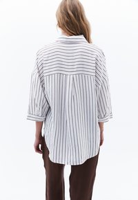 Camicia a righe bianca e marrone, con una vestibilità ampia, con yoke posteriore e maniche arrotolate. Abbinata a pantaloni larghi marroni.