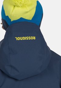 Giacca della marina con collo alto, che mostra il marchio "Rossi" in giallo brillante. Un berretto lavorato a maglia colorato presenta un pompon sulla parte superiore.