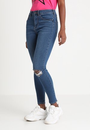 Frau trägt blaue Skinny Jeans mit einem Riss am Knie, ein pinkes Top und weiße Sneaker vor einem einfarbigen Hintergrund.