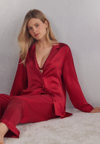 Röd satinpyjamasset med en knapp-up skjorta som har en spetskantad krage och matchande byxor. Slät textur, långa ärmar och avslappnad passform.