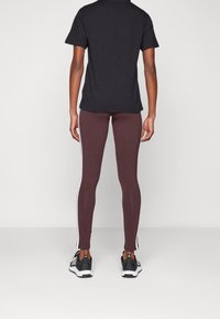 Person som bär mörklila leggings med vita sidostripes, svart t-shirt och svarta Adidas-sneakers, stående mot en enkel bakgrund.