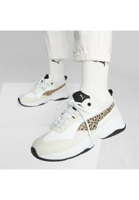Person som bär vita Puma-sneakers med leopardmönster, vita Puma-strumpor och vita uppvikta byxor på en vit yta.
