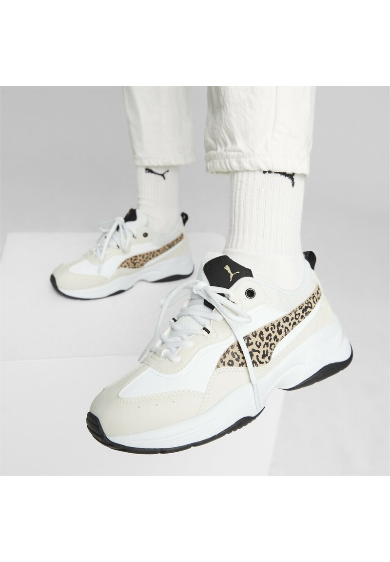 Person som bär vita Puma-sneakers med leopardmönster, vita Puma-strumpor och vita uppvikta byxor på en vit yta.