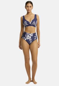 Osoba stoi w pewnej postawie w granatowym, kwiecistym bikini, które składa się z skręconego topu i wysokich majtek, boso na neutralnym tle.