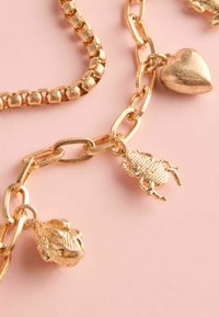 Guldfarvet kædearmbånd med rundede led, teksturerede dyre charms og en hjerteanhæng. Baggrunden er en lyserød overflade.