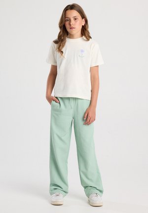 T-shirt en coton blanc avec un graphisme de palmier en violet et vert, associé à un pantalon décontracté texturé vert clair et des baskets blanches.