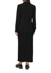 Schwarzes Langarmhemd mit Kragen und strukturiertem Stoff, getragen über einem schwarzen Maxikleid. Das Schuhwerk hat eine klobige Sohle und eine glänzende Oberfläche.