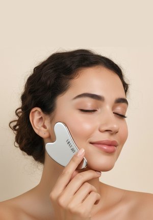 Silver heart-shaped facial massage tool held against the cheek; smooth surface with a logo; model has glowing skin and curly hair.