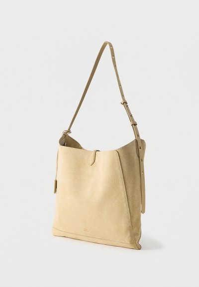 Μπεζ σουέντ τσάντα ώμου με ρυθμιζόμενο λουράκι και μινιμαλιστικό σχεδιασμό, σε όρθια θέση πάνω σε απλό φωτεινό φόντο.