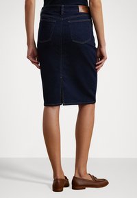 Gonna a tubino in denim scuro con spacco sul retro, accentuata da una toppa in pelle marrone e cuciture a contrasto. Indossata con mocassini marroni.