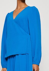 Blue wrap blouse with long sleeves, featuring a textured fabric and deep V-neckline. Puffed sleeves end with elastic cuffs.