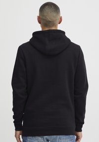 Sudadera negra con una textura suave, capucha con cordón y puños acanalados. La parte posterior presenta un diseño sin costuras, combinada con jeans azul desgastados.