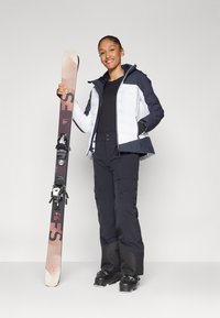 Skiausrüstung mit einer schwarz-weißen isolierten Jacke, schwarzen Hosen und schwarzen Skischuhen, die einen hellen Ski mit Bindungen hält.