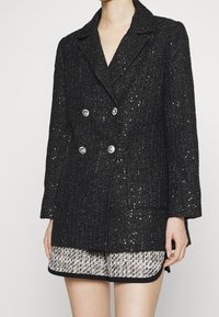 Černý tvídový dvouřadý blazer se stříbrnými knoflíky, s texturovaným povrchem a klopovými límci, v kombinaci s světlými patterned šortkami.