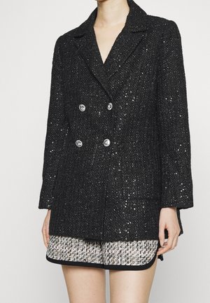 Blazer de tweed negro de doble botonadura con botones plateados, que presenta un acabado texturizado y solapas con muescas, combinado con pantalones cortos de color claro y estampados.