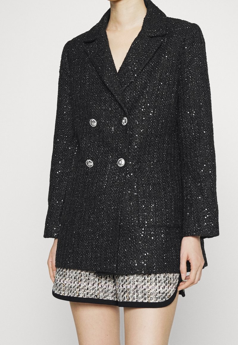 Černý tvídový dvouřadý blazer se stříbrnými knoflíky, s texturovaným povrchem a klopovými límci, v kombinaci s světlými patterned šortkami.