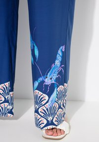 Námořnické modré kalhoty s potiskem humra, opatřené modrými a růžovými akcenty. Spodní lem má bílý vzor mušlí, což vytváří kontrastní designový prvek.
