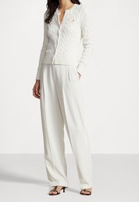 Cárdigan blanco de punto con un cuello redondo y un pequeño logotipo dorado, combinado con pantalones blancos holgados y tacones negros con tiras.