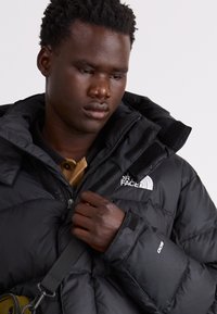 Svart puffjacka med hög krage, med en vit logotyp på bröstet och kardborreknäppning vid ärmarna. Bär en brun skjorta under.