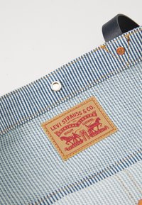 Kék és fehér csíkos farmer anyagú táska barna bőr füllel, valamint egy bézs címkével, amelyen piros grafikák és szöveg található a Levi Strauss & Co. től.