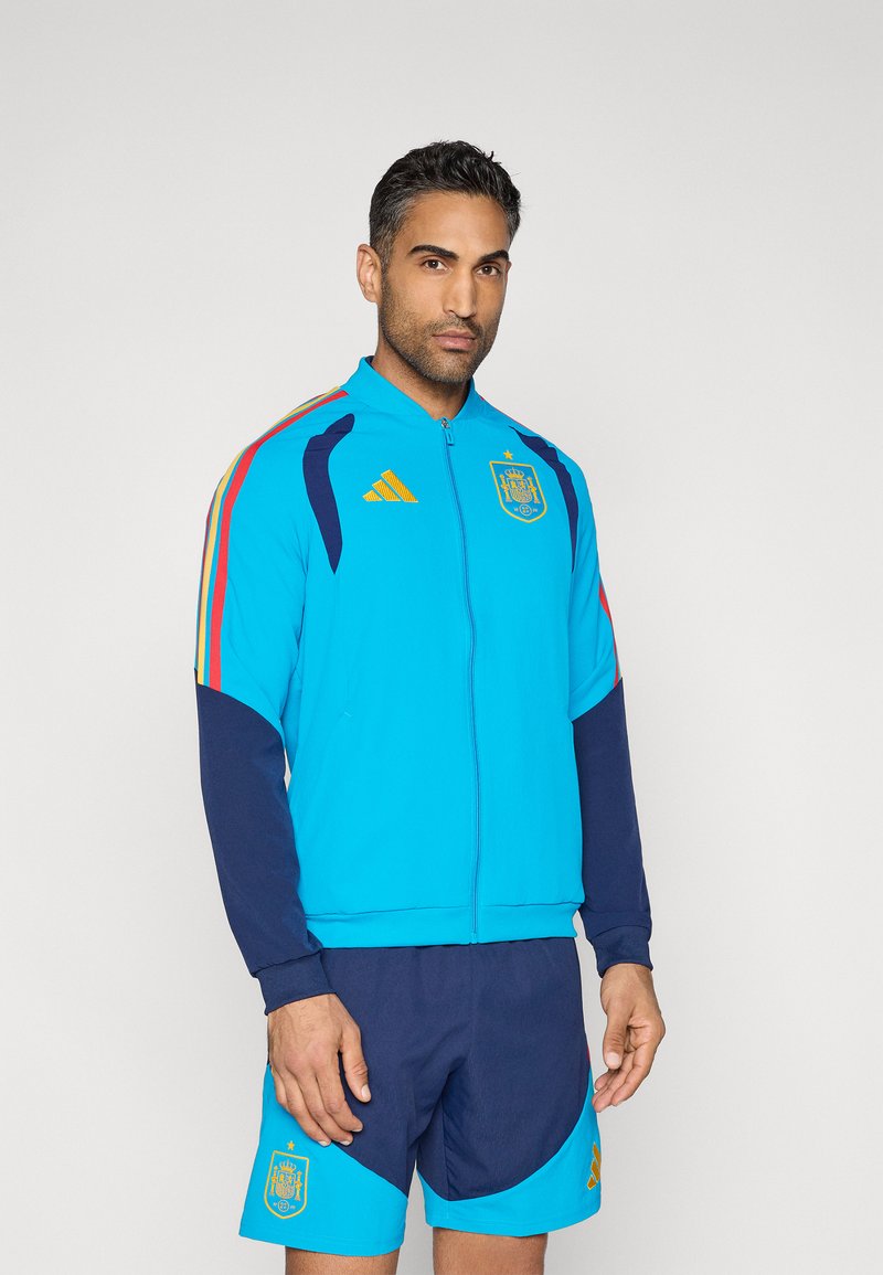 Kék dzseki cipzárral, tengerészkék ujjakkal és piros részletekkel. Spanyolország címerével és Adidas logóval díszítve. Tengerészkék rövidnadrággal párosítva. Sima textúrájú.