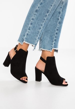 Zwarte suède open-toe slingback hakken gedragen met lichtblauwe jeans met rafelige zoom op een witte reflecterende vloer.