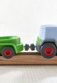 Blauer Spielzeugtraktor aus Kunststoff mit roten Rädern, gekoppelt an einen grünen Anhänger. Auffälliges Rad- und Verbindungsdesign auf einer Holzschiene.