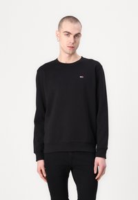 Sudadera negra con cuello redondo, mangas largas, puños y dobladillo acanalados, que presenta un pequeño logo en el pecho. La tela parece suave y lisa.