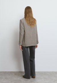 Blazer de cuadros en beige y marrón con mangas largas, combinado con pantalones de pierna ancha en gris oscuro. La tela parece tener textura y un ajuste entallado.