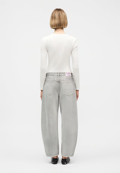 Γυναίκα με κοντά ξανθά μαλλιά, φορώντας λευκή μάλλινη μπλούζα με μακριά μανίκια και άνετο ανοιχτό γκρι τζιν, στέκεται με την πλάτη προς τα εμπρός σε απλό φόντο.