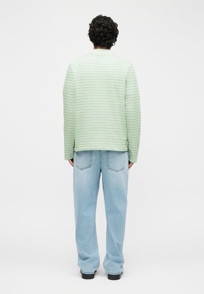 Άνδρας με σγουρά μαλλιά στέκεται πλάτη, φορώντας μακρυμάνικο πουκάμισο με πράσινες και λευκές ρίγες και φαρδιά ανοιχτό μπλε τζιν με μαύρα παπούτσια.