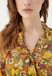 Blusa floreale gialla, con vivaci motivi arancioni, blu e verdi. Il tessuto sembra leggero con un design a collare a scollatura.