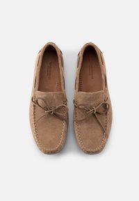 Ζευγάρι ανοιχτόχρωμων καφέ σουέντ μοκασίνια με φιόγκο, ορατή ραφή και επενδυμένα πέλματα σε απλό λευκό φόντο.