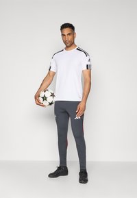 T-shirt de sport blanche avec des rayures noires sur les épaules, associée à des leggings gris avec des accents rouges sur les côtés et un logo Adidas. Tenant un ballon de soccer.