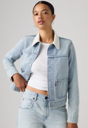 Jeune femme portant une veste en jean bleu clair avec un col blanc, un top court blanc et un jean assorti, posant avec la main près de la taille.