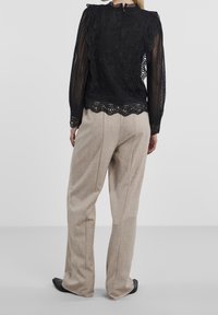 Haut en dentelle noire à manches longues, présentant un motif élaboré et un ourlet en dents de scie. Associé à un pantalon en tissu beige et des chaussures noires.